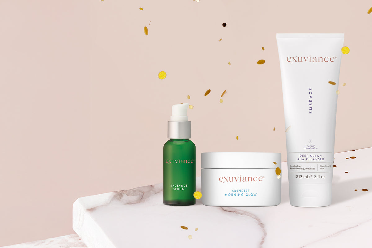 Three Exuviance products and gold christmas ornaments on a marble counter top against a light beige background with gold glitter flying in the air.
