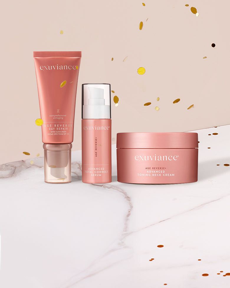 Three Exuviance AGE REVERSE+ products on a marble counter top against a light beige background with gold glitter flying in the air.
