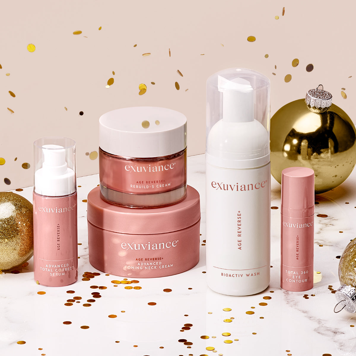 The Exuviance AGE REVERSE+ collection and gold christmas ornaments on a marble counter top against a light beige background with gold glitter flying in the air.