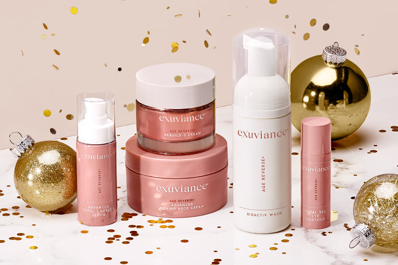 The Exuviance AGE REVERSE+ collection and gold christmas ornaments on a marble counter top against a light beige background with gold glitter flying in the air.