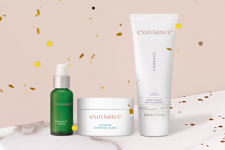 Three Exuviance products and gold christmas ornaments on a marble counter top against a light beige background with gold glitter flying in the air.