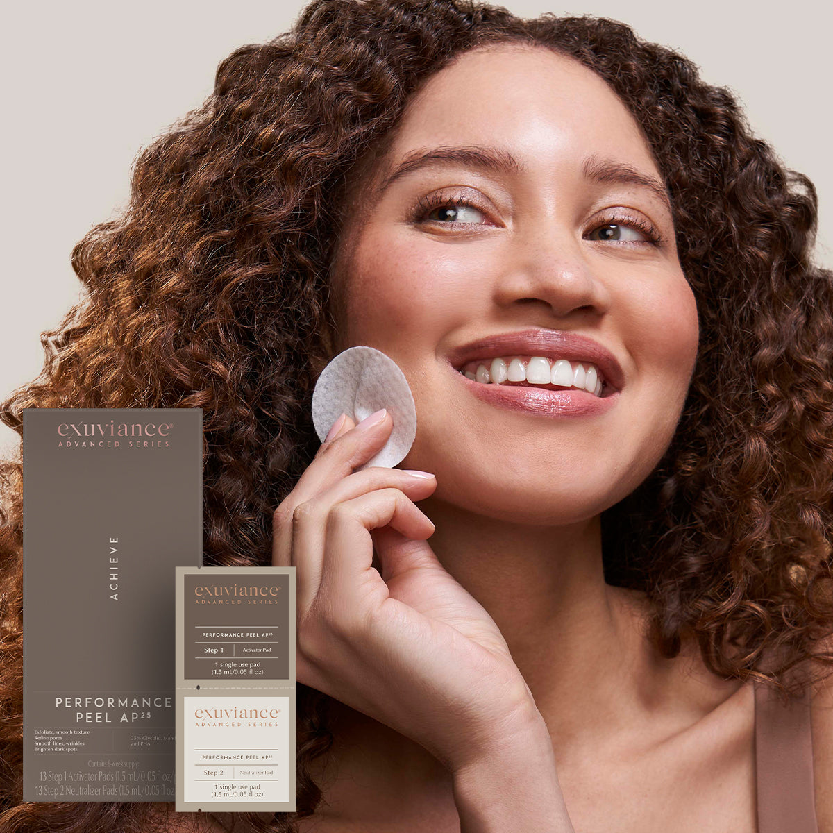 A woman with brown curly hair wearing a tank top and smiling holding a Performance Peel AP 25 peel pad to her face, against a light beige background.