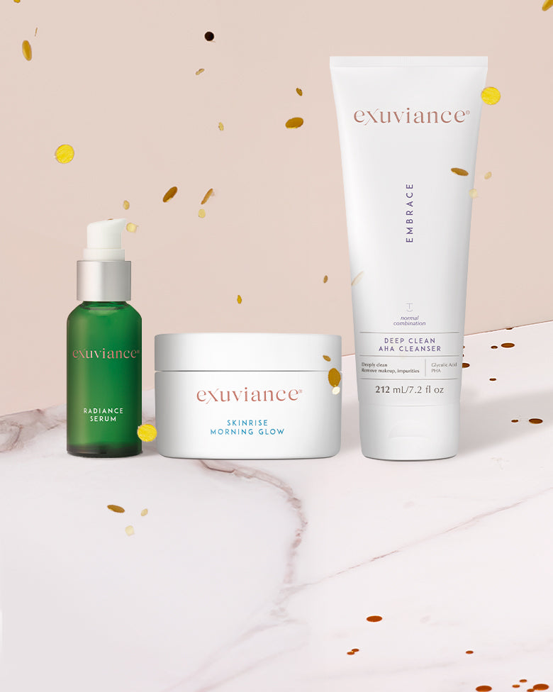 Three Exuviance products and gold christmas ornaments on a marble counter top against a light beige background with gold glitter flying in the air.
