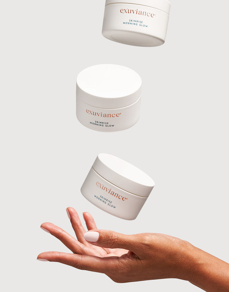 Three bottles of Exuviance SkinRise Morning Glow floating on a white background above a woman's open palm. 