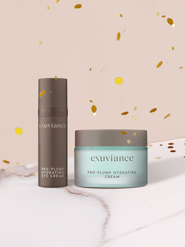 Exuviance Pro-Plump Hydrating Eye Cream and Pro-Plump Hydrating Cream on a marble countertop, against a light beige background with glitter flying in the air.