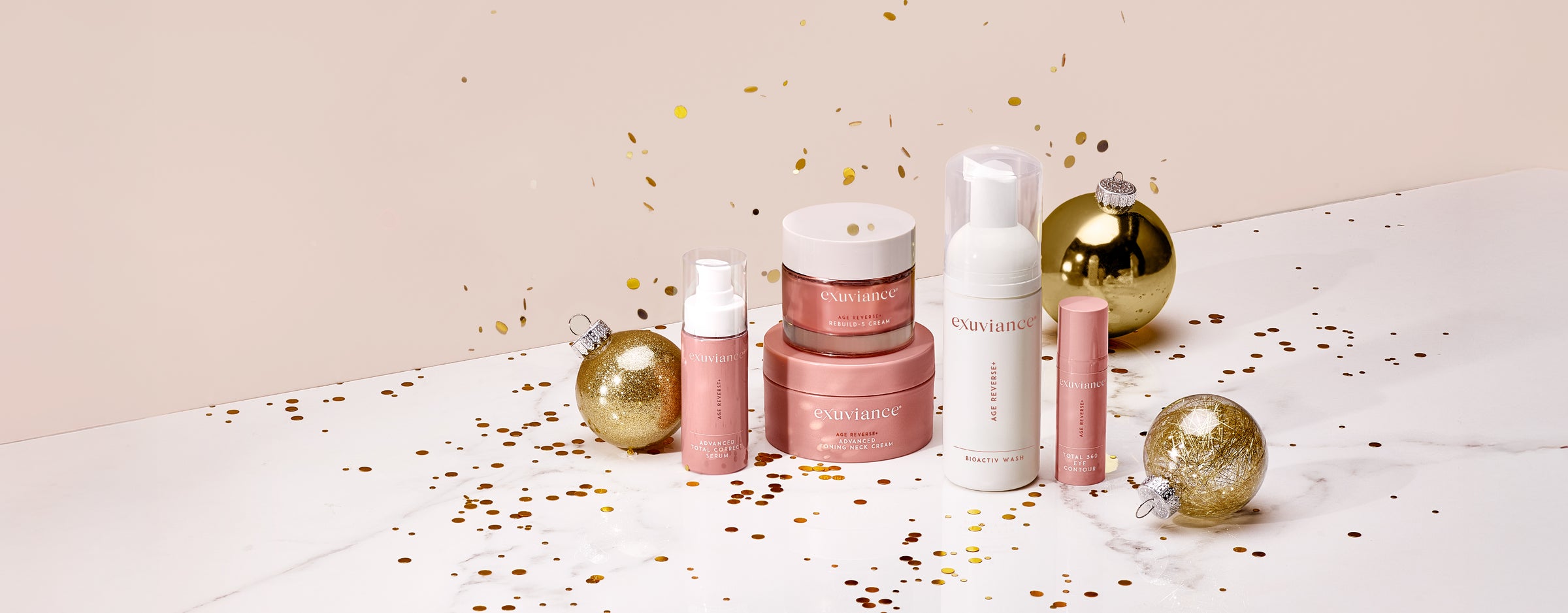 The Exuviance AGE REVERSE+ collection and gold christmas ornaments on a marble counter top against a light beige background with gold glitter flying in the air.