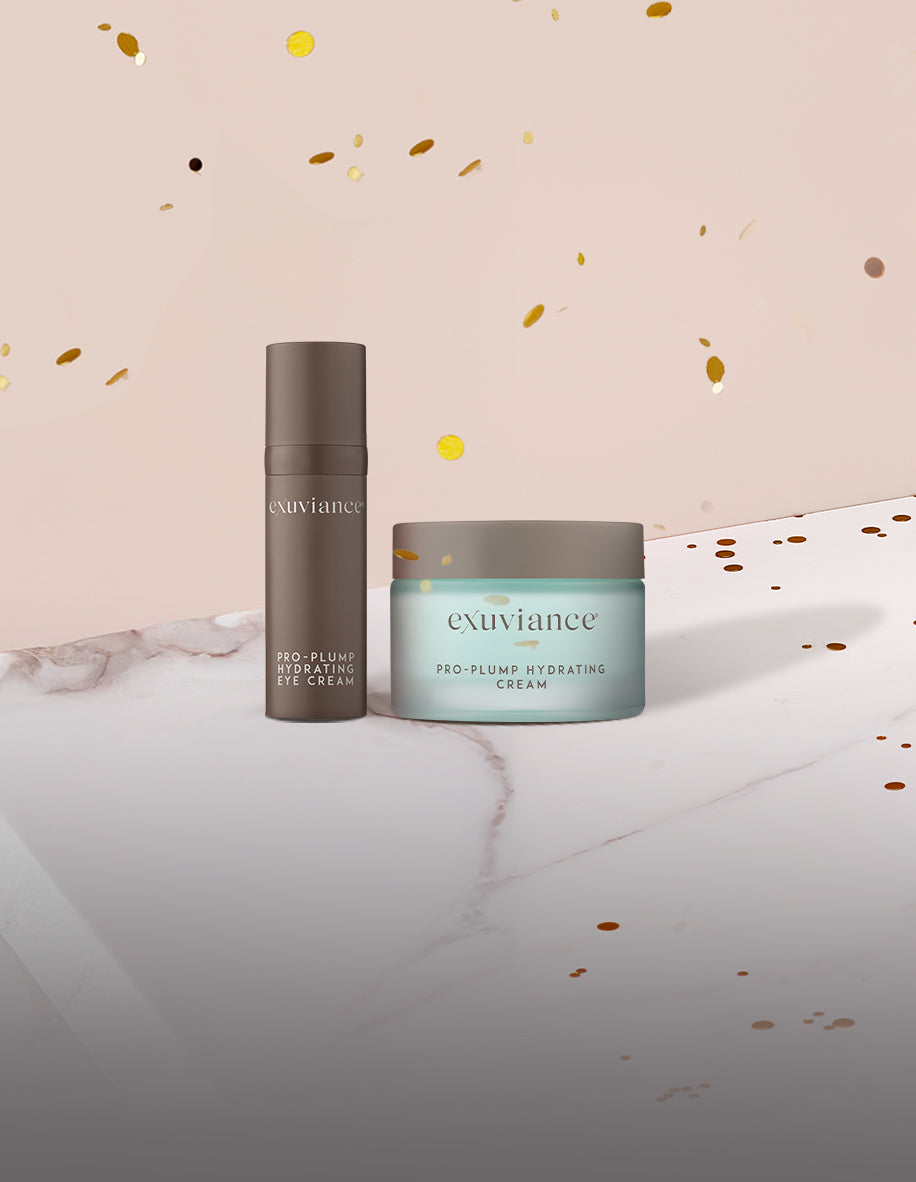 Exuviance Pro-Plump Hydrating Cream and Eye Cream and gold christmas ornaments on a marble counter top against a light beige background with gold glitter flying in the air.