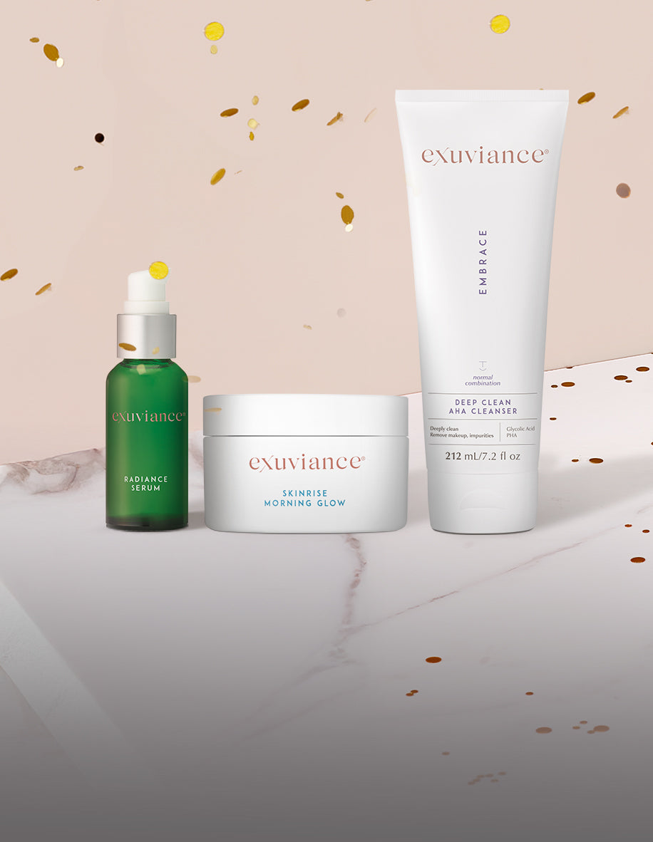 Three Exuviance products and gold christmas ornaments on a marble counter top against a light beige background with gold glitter flying in the air.