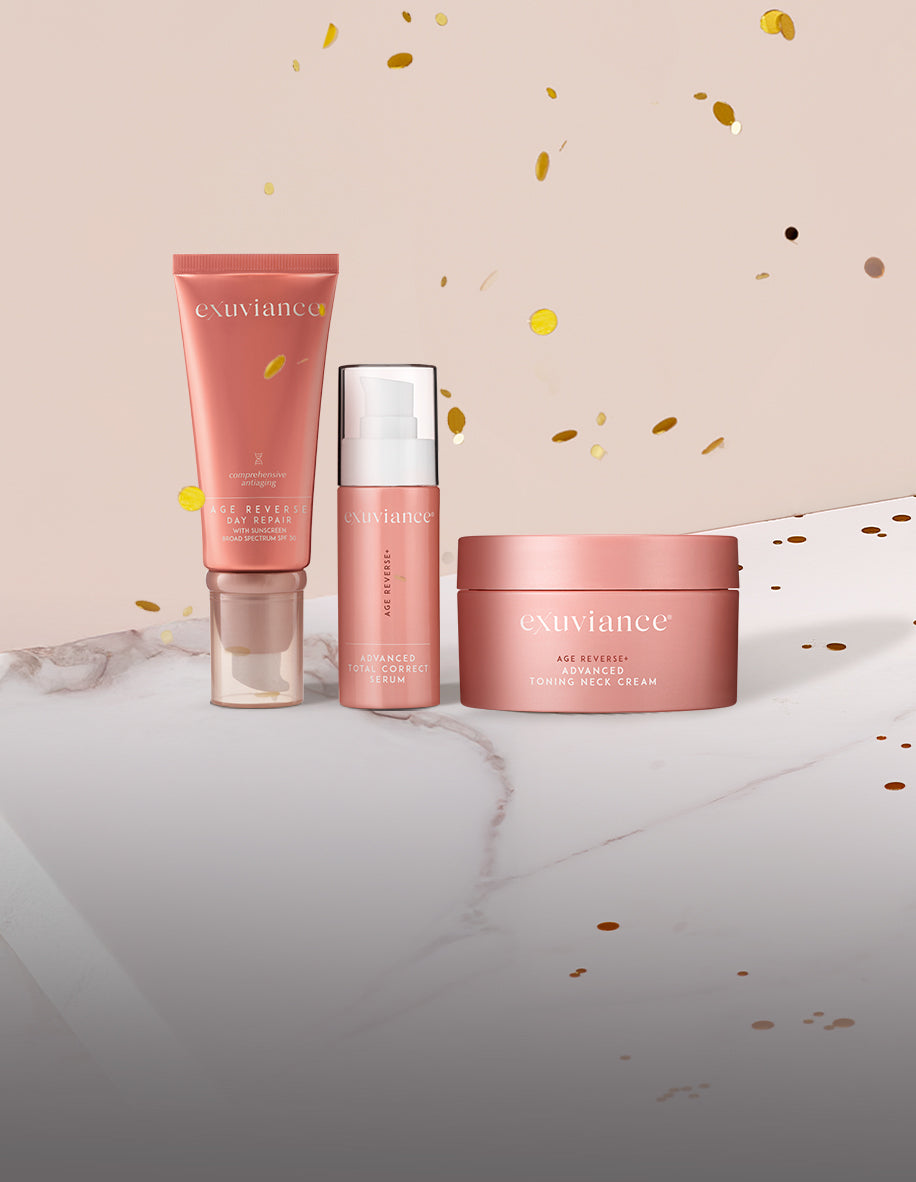 Three Exuviance AGE REVERSE+ products and gold christmas ornaments on a marble counter top against a light beige background with gold glitter flying in the air.