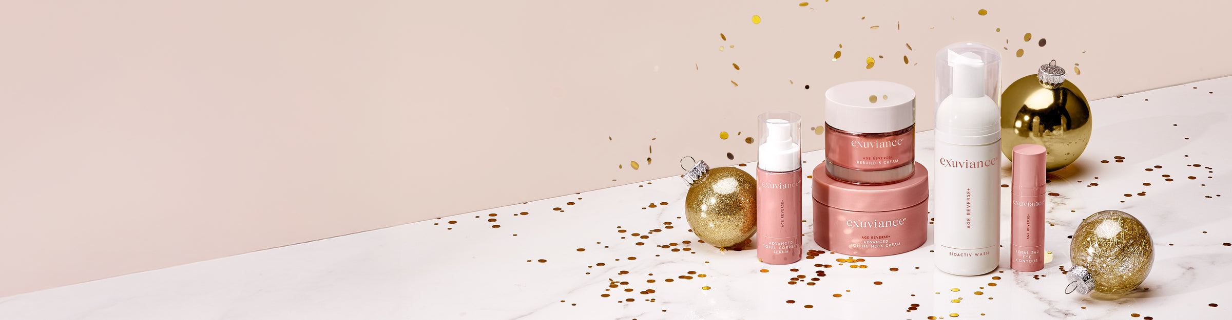The Exuviance AGE REVERSE+ collection and gold christmas ornaments on a marble counter top against a light beige background with gold glitter flying in the air.