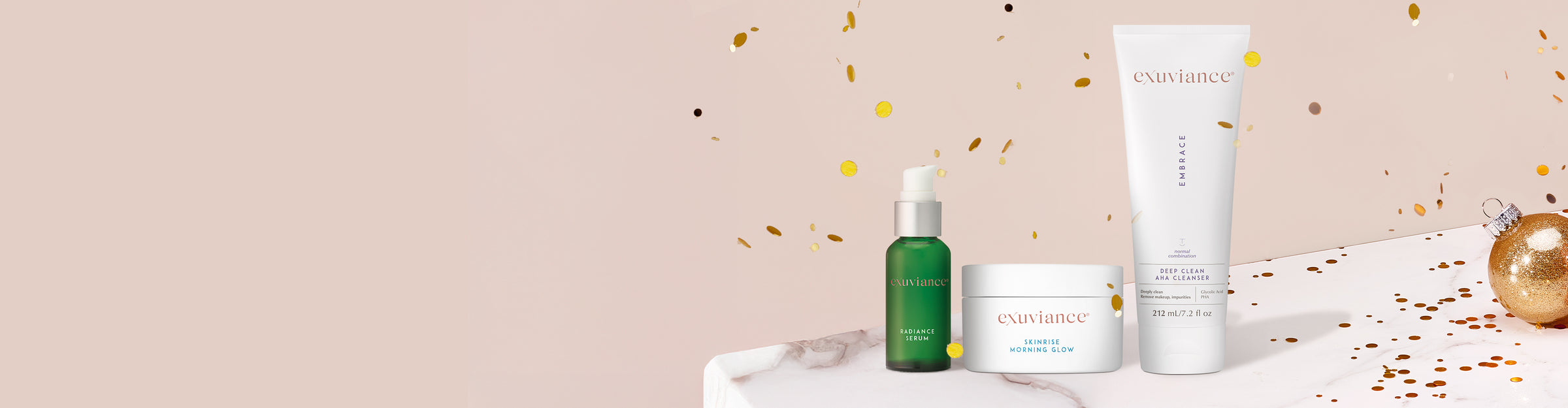 Three Exuviance products and gold christmas ornaments on a marble counter top against a light beige background with gold glitter flying in the air.