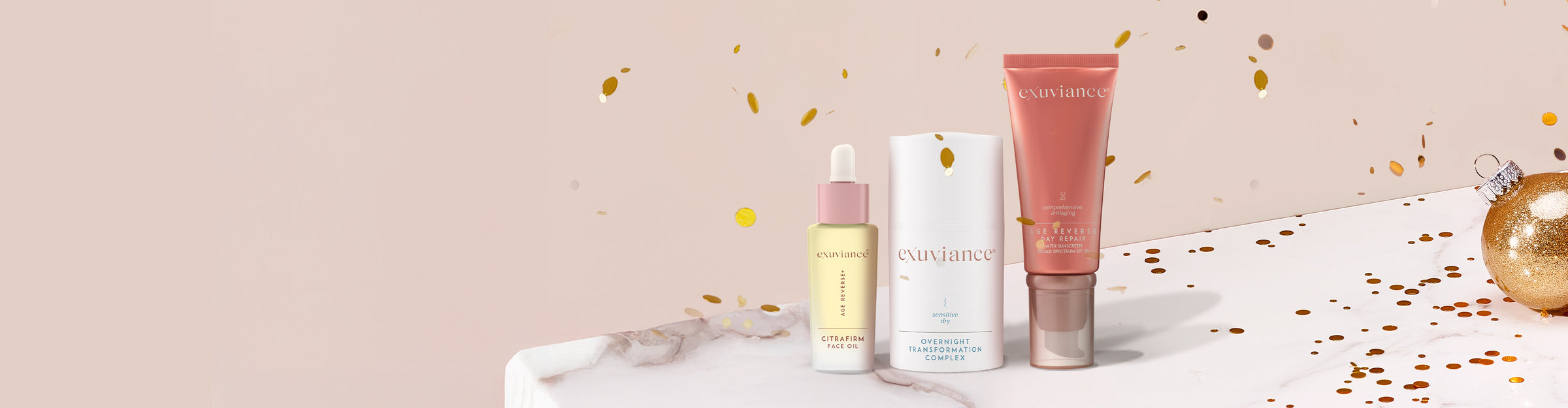 Three Exuviance products and gold christmas ornaments on a marble counter top against a light beige background with gold glitter flying in the air.