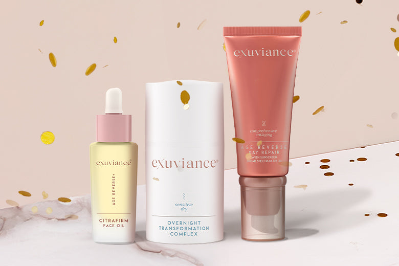 Three Exuviance products and gold christmas ornaments on a marble counter top against a light beige background with gold glitter flying in the air.