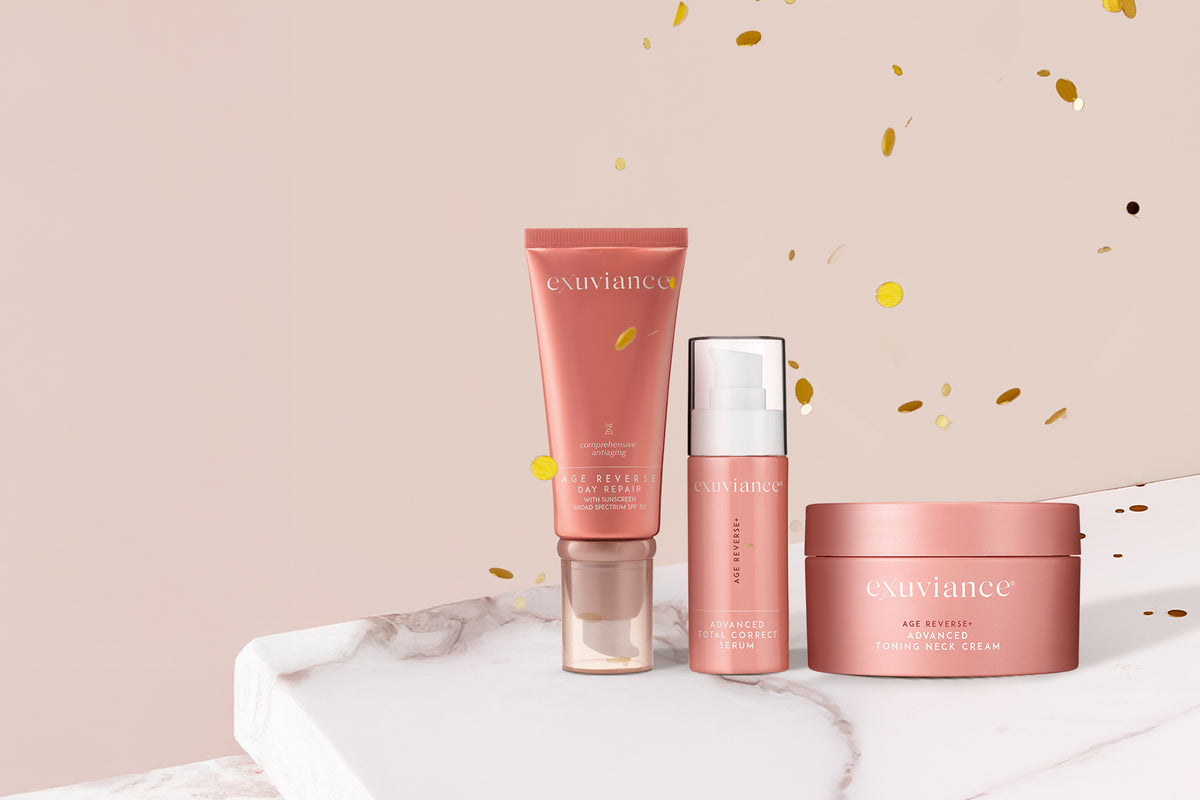 Three Exuviance AGE REVERSE+ products on a marble counter top against a light beige background with gold glitter flying in the air.